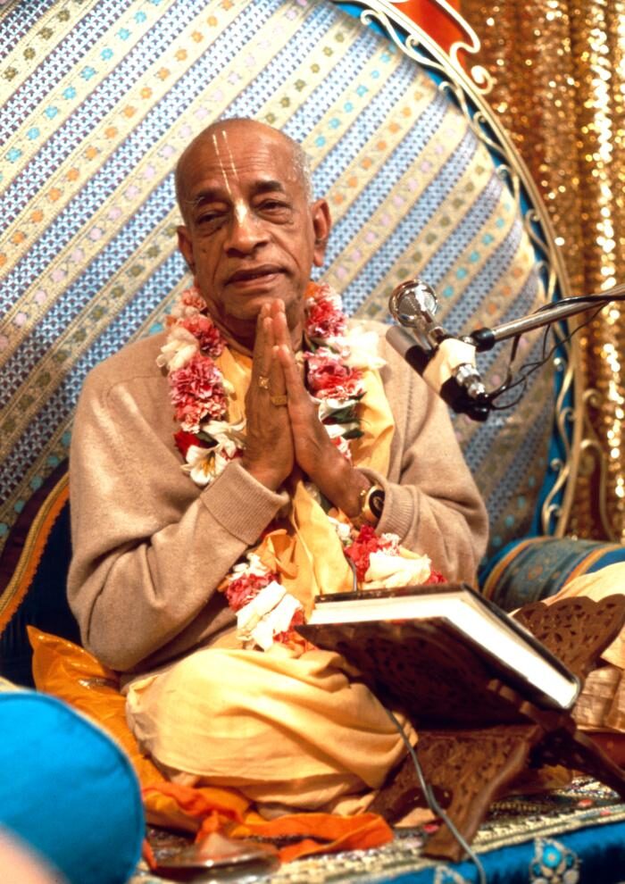Srila Prabhupada seated at his vyasasana with hands folded, a book stand and microphone beside him.