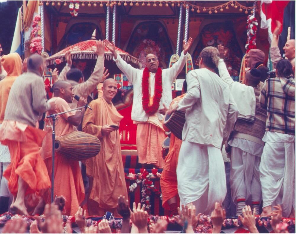 Srila Prabhupada dancing with arms raised atop a Ratha-yatra cart, surrounded by disciples playing mridanga, with a crowd cheering below.
